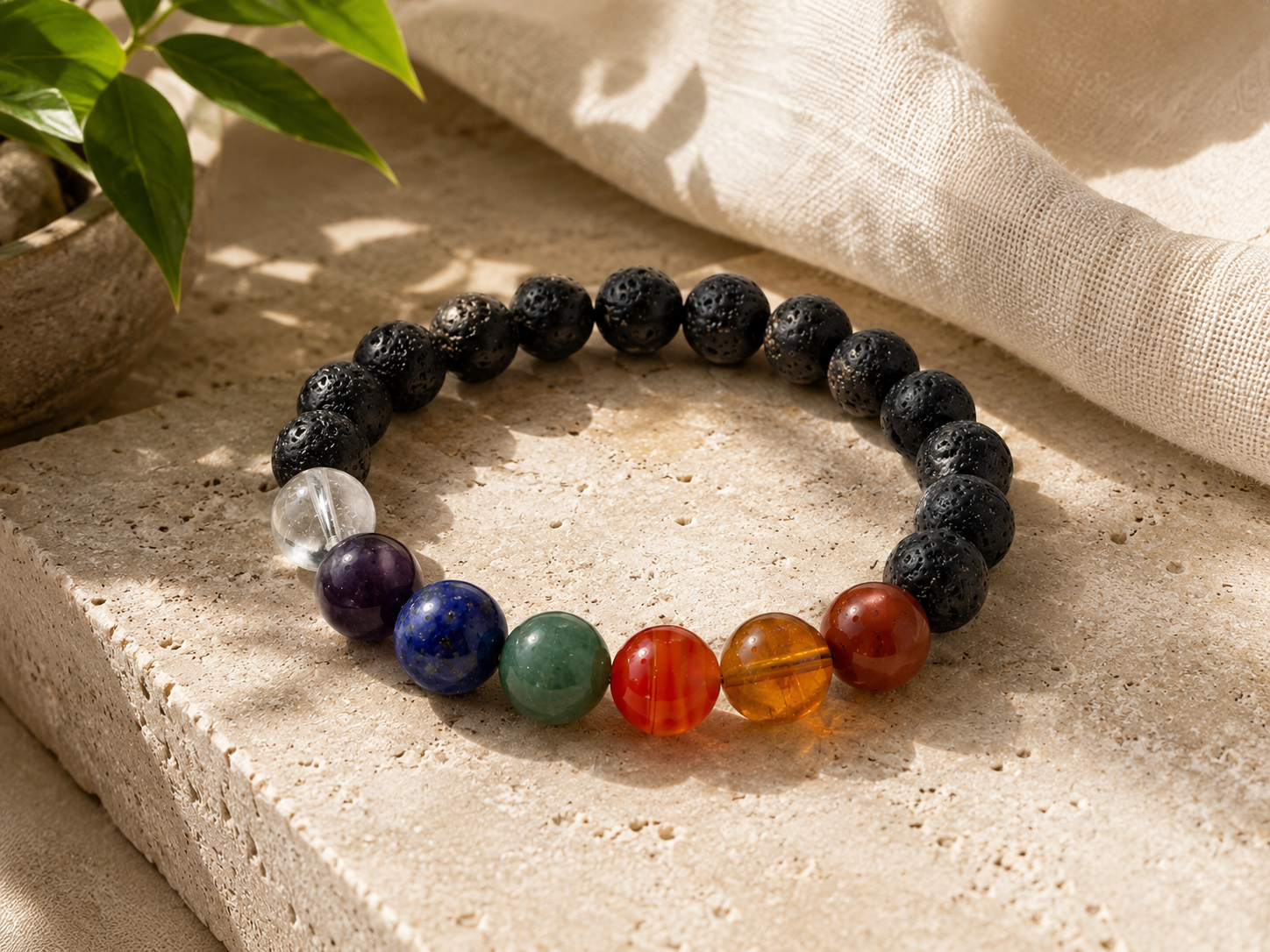 Lava Stone 7 Chakra Bracelet resting on a pale stone surface with linen and a green plant