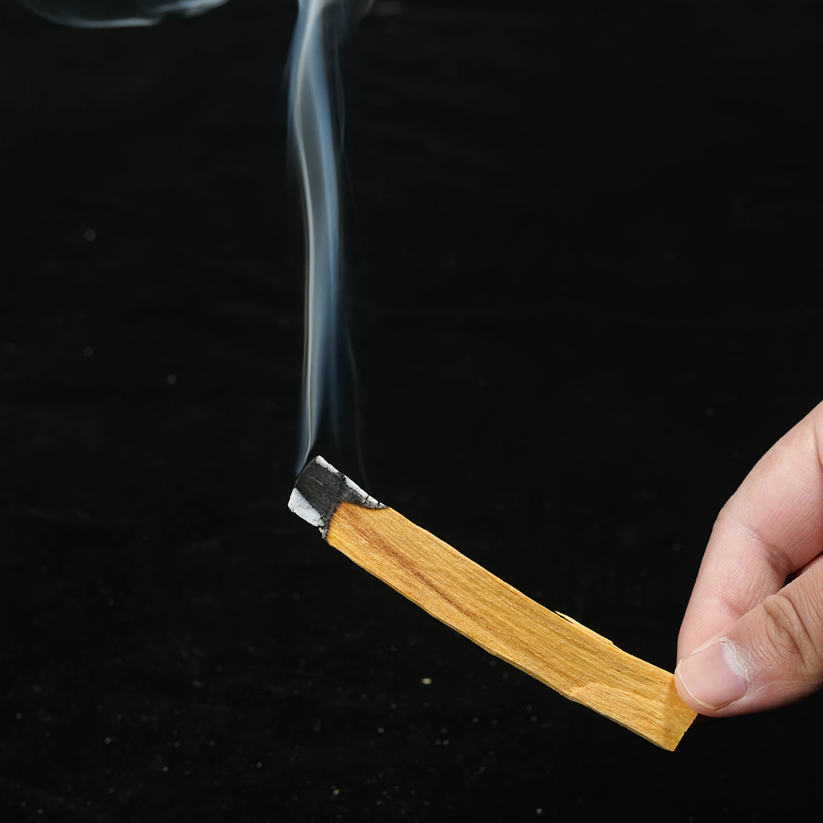 A hand holding a lit palo santo stick with a thin ribbon of smoke rising
